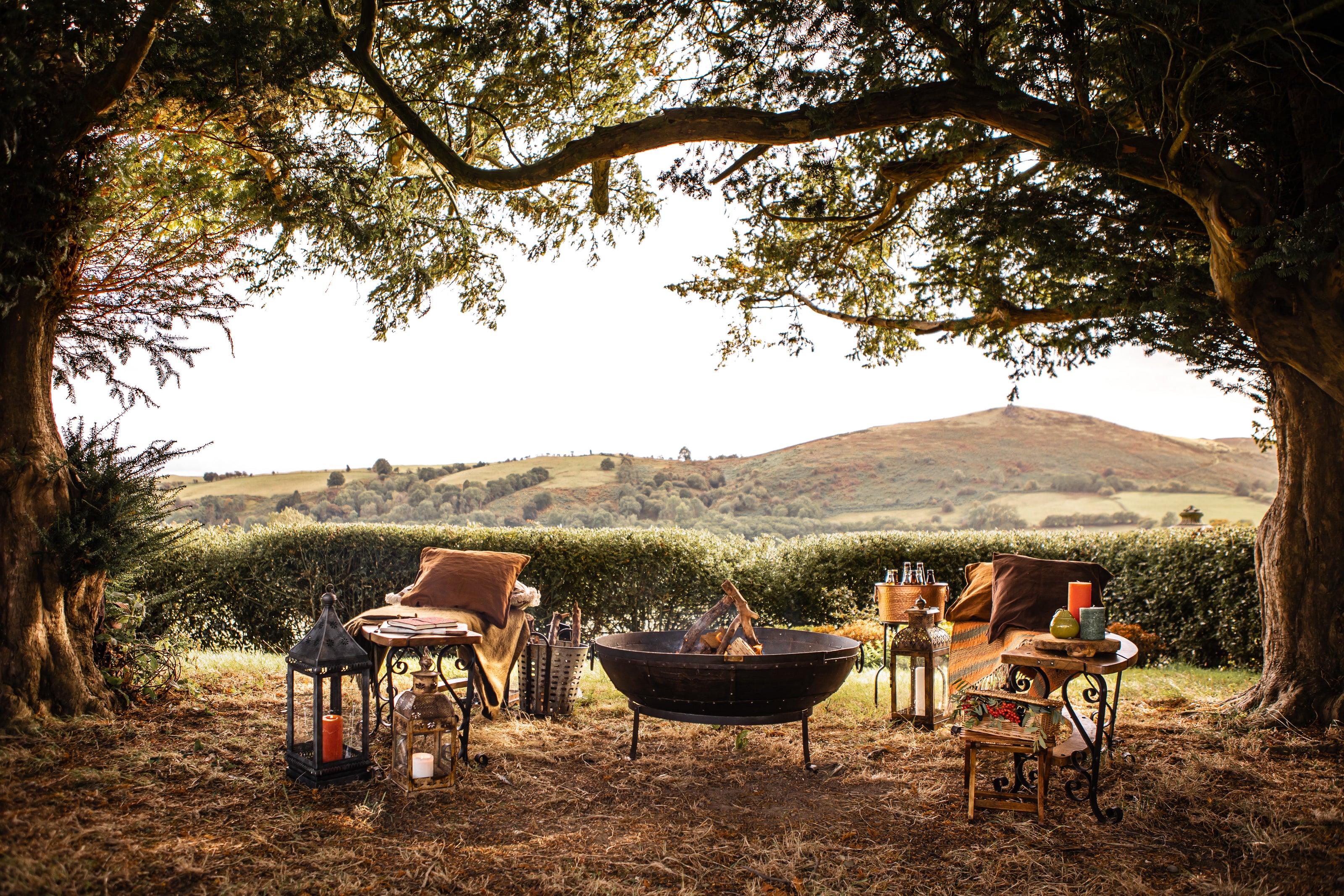 Outdoor setting with a kadai fire pit, chairs, and lanterns under large trees with a scenic background.