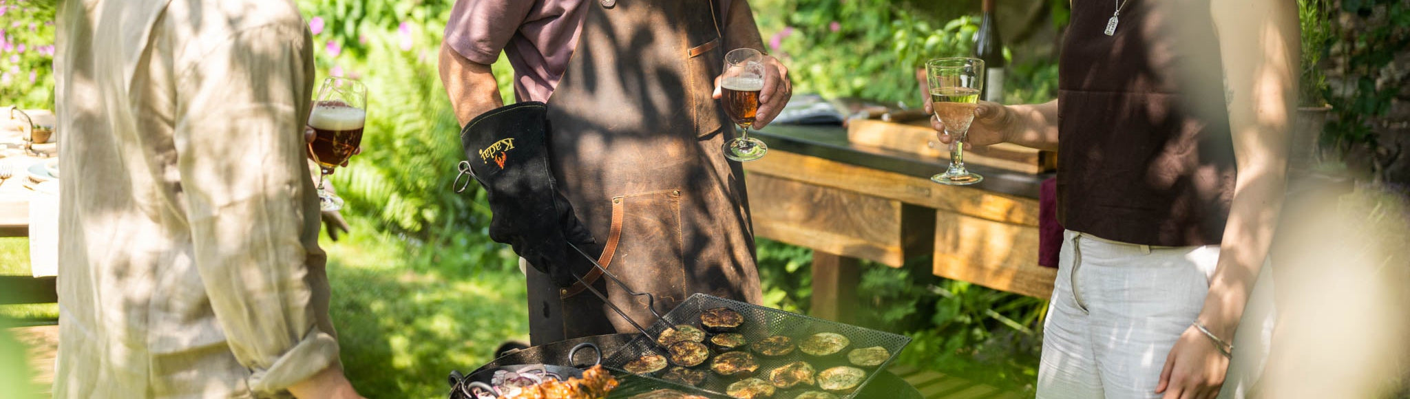 Three people grilling outdoors in a garden setting on a Kadai charcoal barbecue