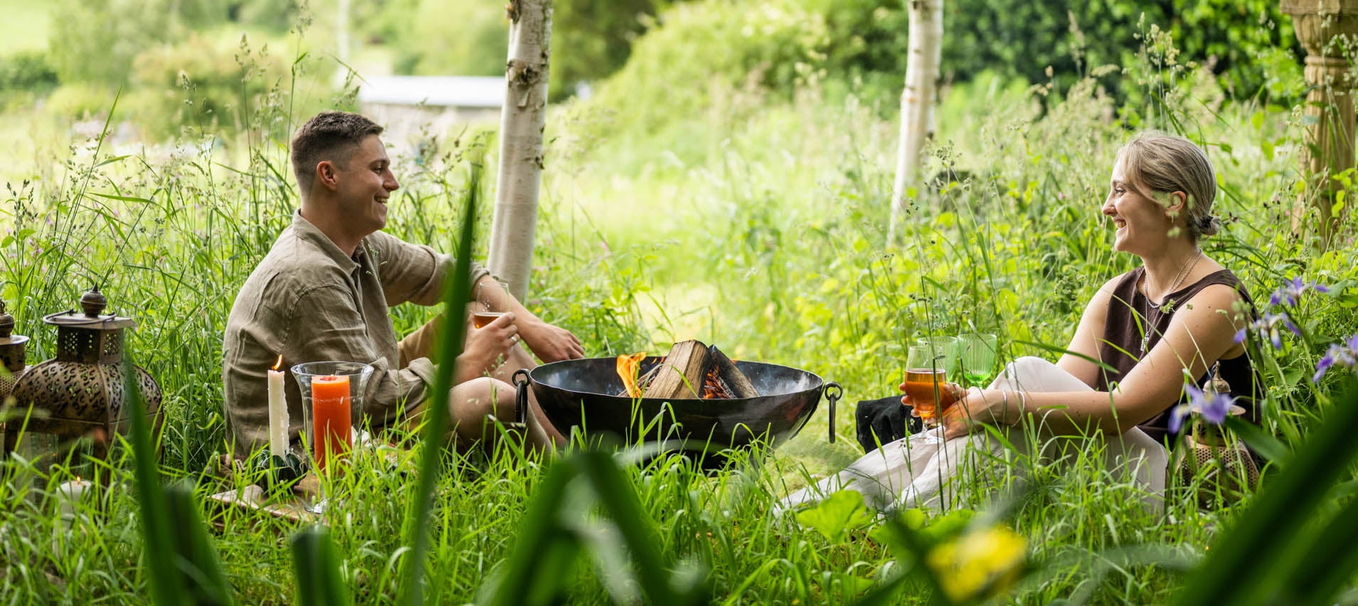 Two people sitting by a Kadai Fire Pit in a natural setting with drinks and a grill.