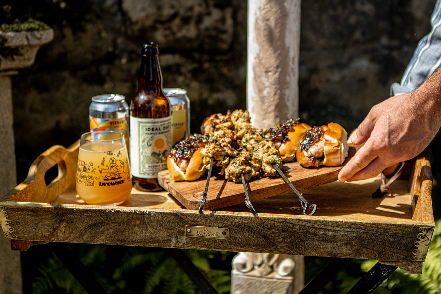 A wooden serving tray outdoors holds several small grilled kebabs topped with a chunky herb sauce, skewers, a glass of beer, a beer bottle, and a can. A person's hand is reaching toward the food, with a stone wall and garden background.