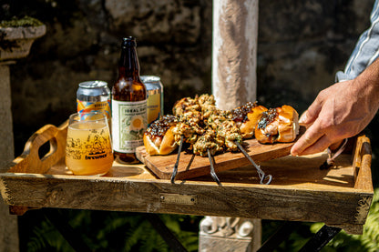 A wooden serving tray outdoors holds several small grilled kebabs topped with a chunky herb sauce, skewers, a glass of beer, a beer bottle, and a can. A person's hand is reaching toward the food, with a stone wall and garden background.