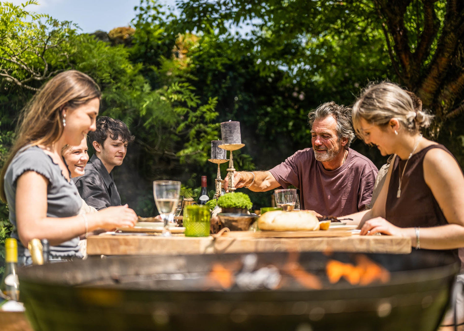 Group of people enjoying a meal around an outdoor fire pit with drinks and food.