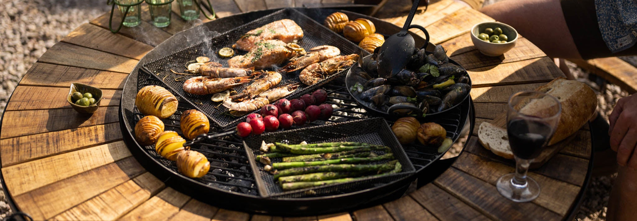 Outdoor setting with a wooden firebowl table featuring a grill pan with food, wine glasses, and people around.