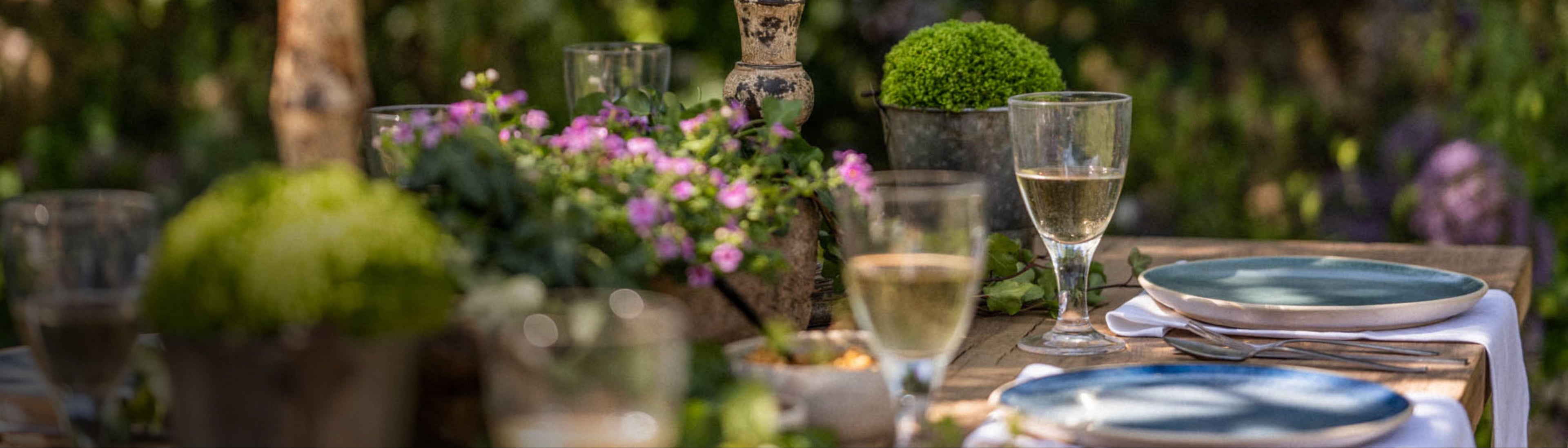 Outdoor table setting with wine glasses, plates, and flowers in a garden.