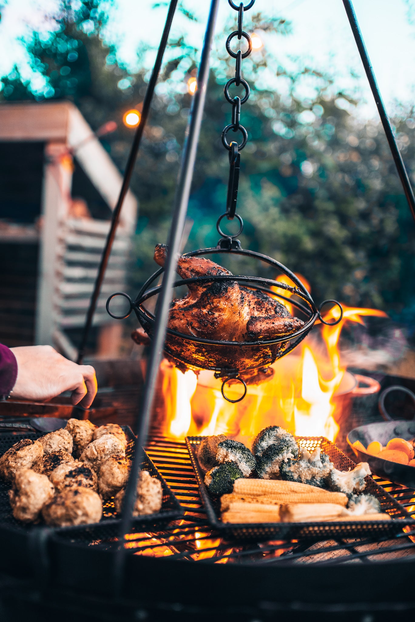 A hanging metal basket suspends over an open fire, cooking a whole roasted chicken outdoors. Below the chicken, grates hold broccoli, corn on the cob, and other grilled items, with bright flames visible in the background.