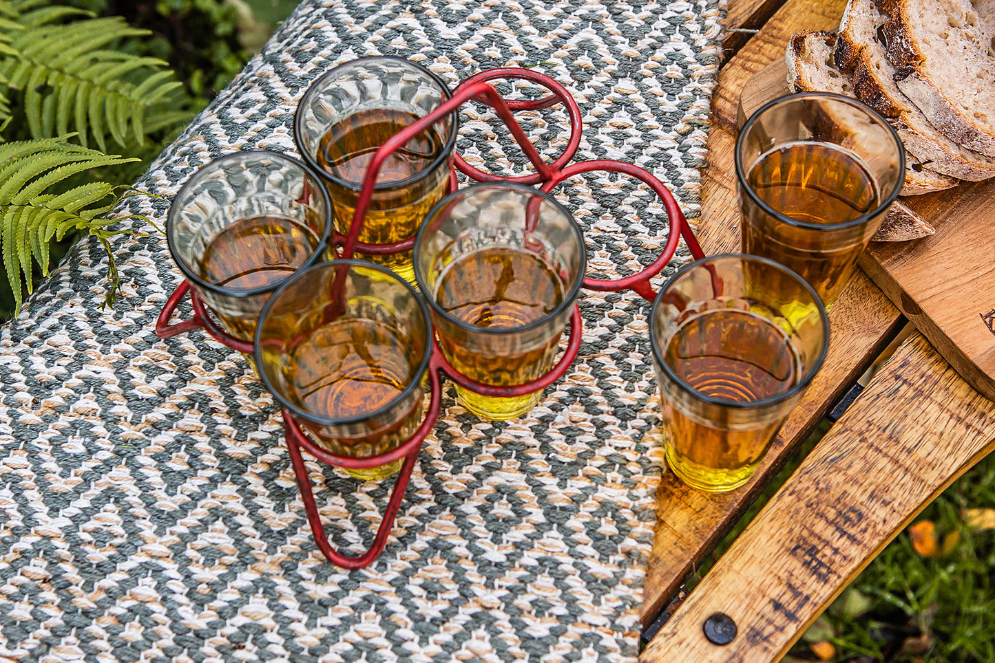 Glasses of beer in a red metal carrier on a patterned tablecloth next to a wooden bench outdoors.
