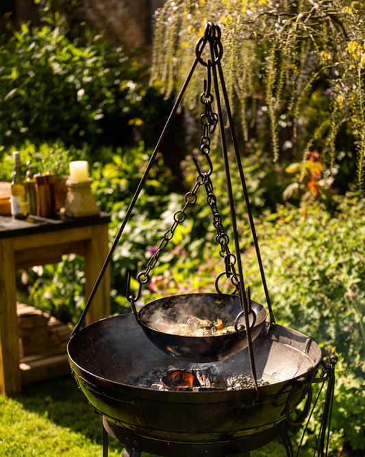 Cooking bowl suspended over a Kadai firebowl barbecue on a hanging tripod stand in an outdoor garden cooking setting