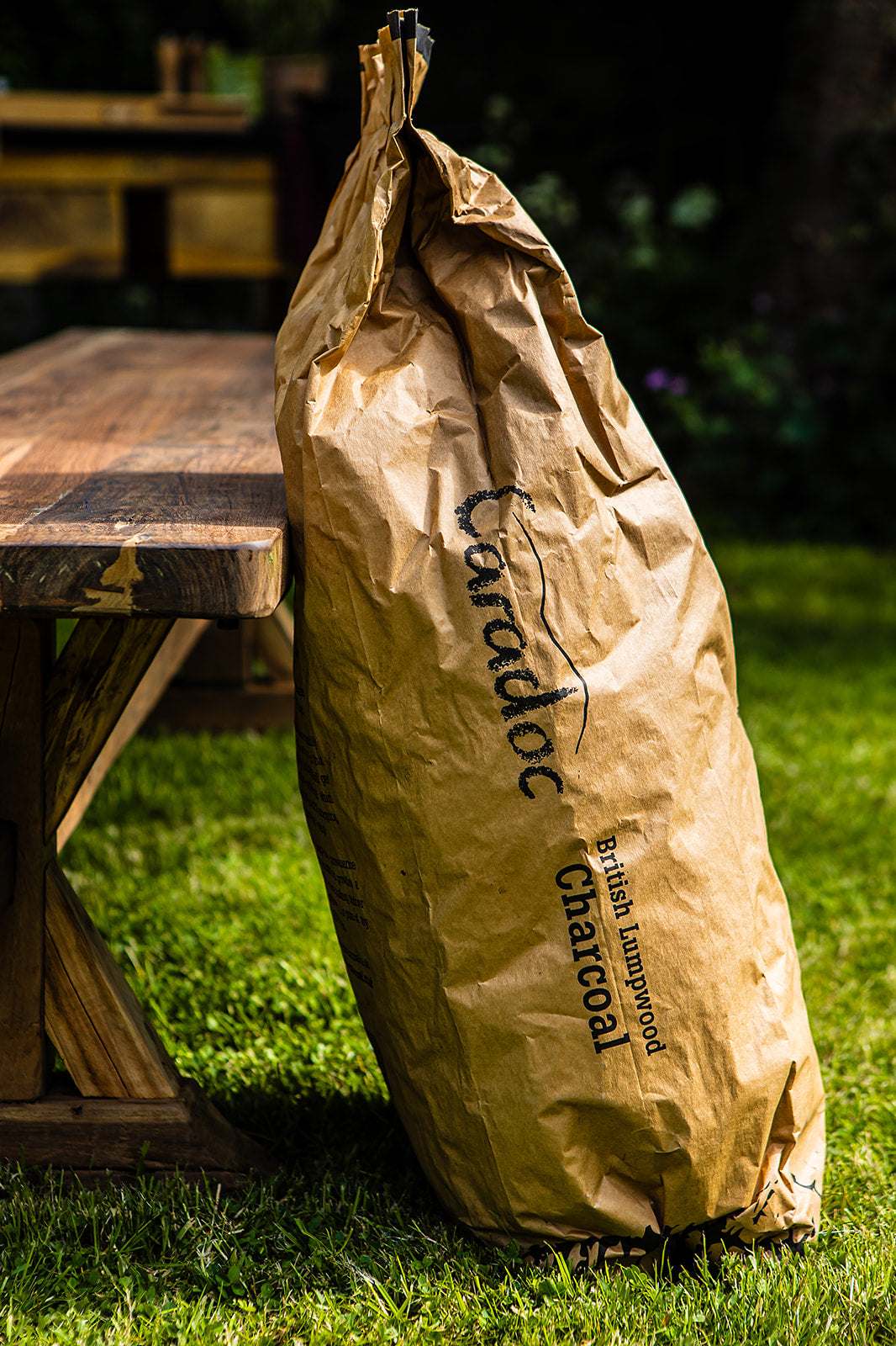 A large brown paper bag of Caradoc British Lumpwood Charcoal sits on a green grass lawn next to a rustic wooden picnic table in an outdoor backyard setting.