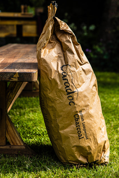 A large brown paper bag of Caradoc British Lumpwood Charcoal sits on a green grass lawn next to a rustic wooden picnic table in an outdoor backyard setting.
