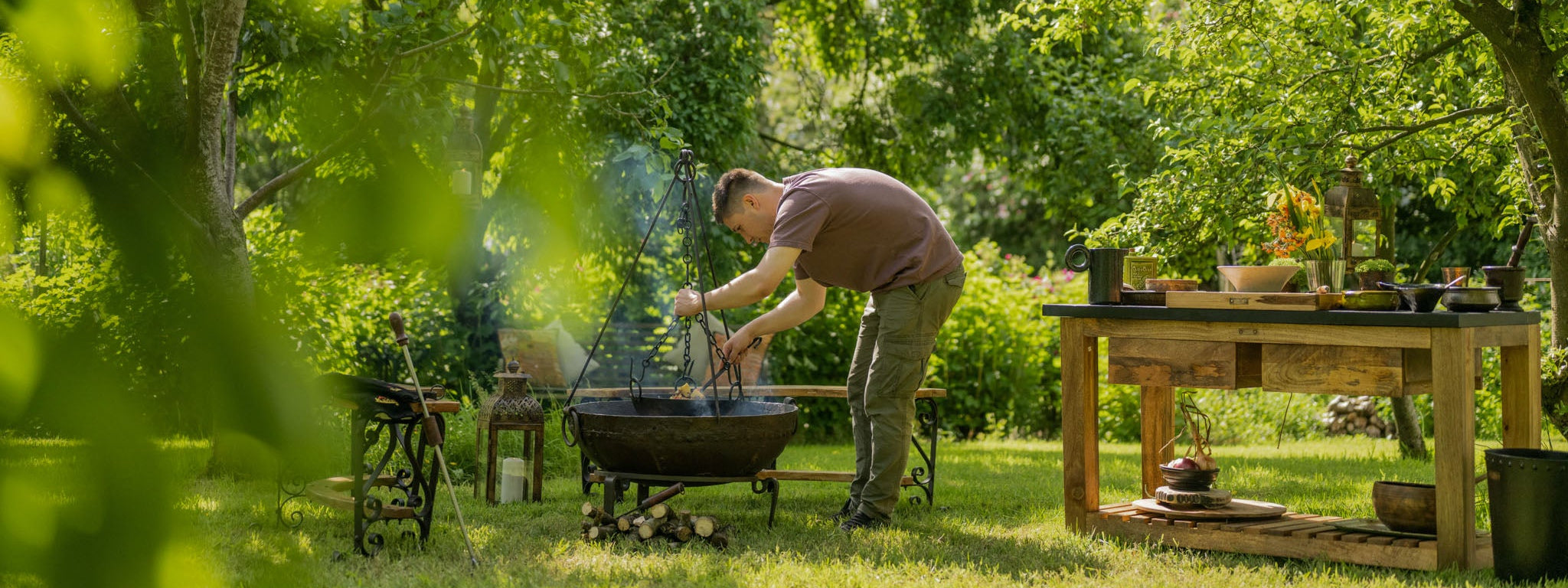 Man tending a live-fire cooking setup in a lush garden, adjusting a hanging tripod grill over a large fire bowl beside a rustic wooden outdoor prep table surrounded by trees and greenery.