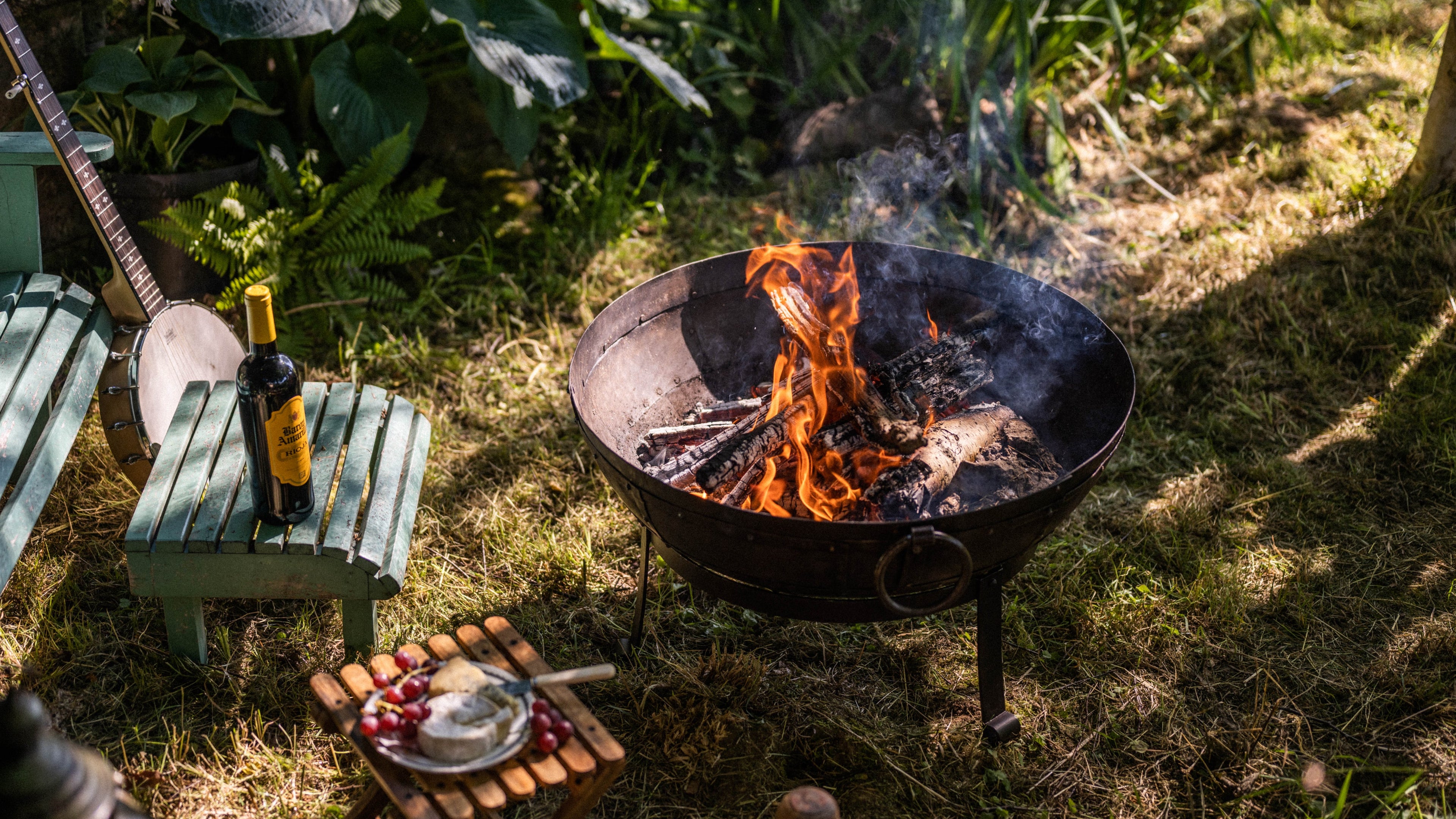 Fire pit with flames and smoke in a garden setting with a bottle and fruits on a small table.