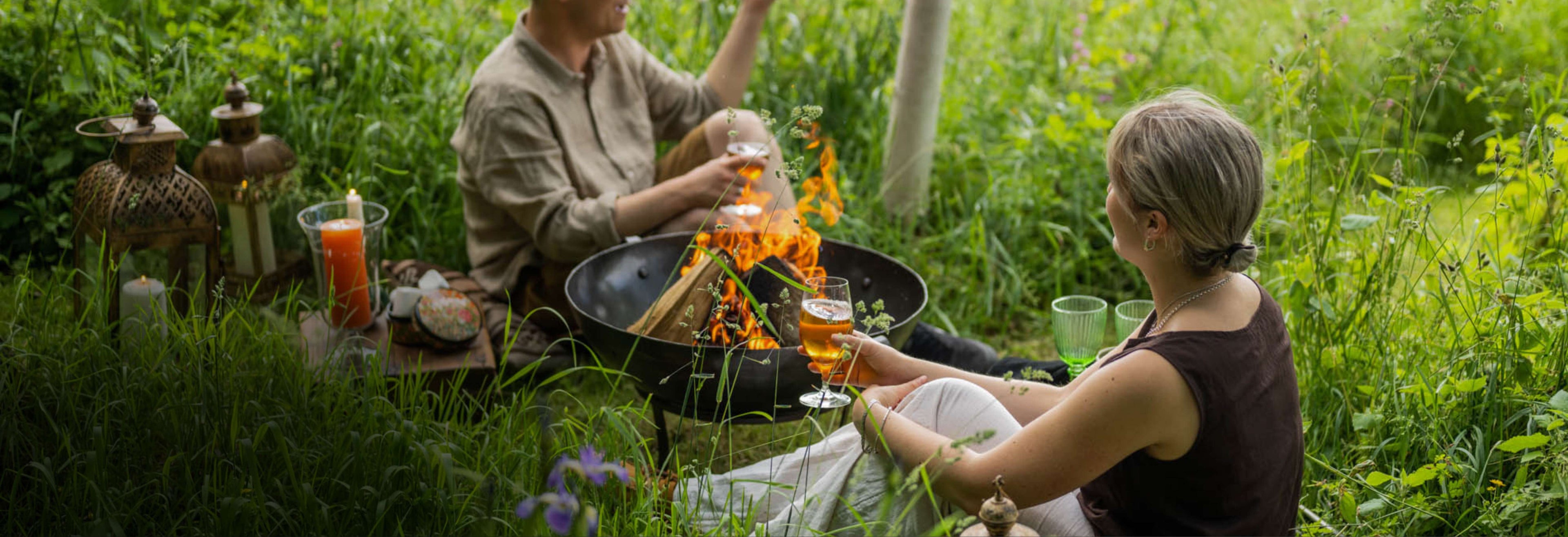 
Two people enjoying a Fire Pit in a grassy field with fireside accessories and drinks
