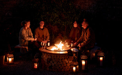 Group of friends gathered around a glowing Kadai firebowl table at night, enjoying drinks and conversation beside lanterns and an open fire in a cozy outdoor garden setting.