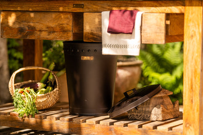 Black BBQ bin with removable lid stored beneath a wooden outdoor kitchen station beside fresh vegetables and barbecue accessories