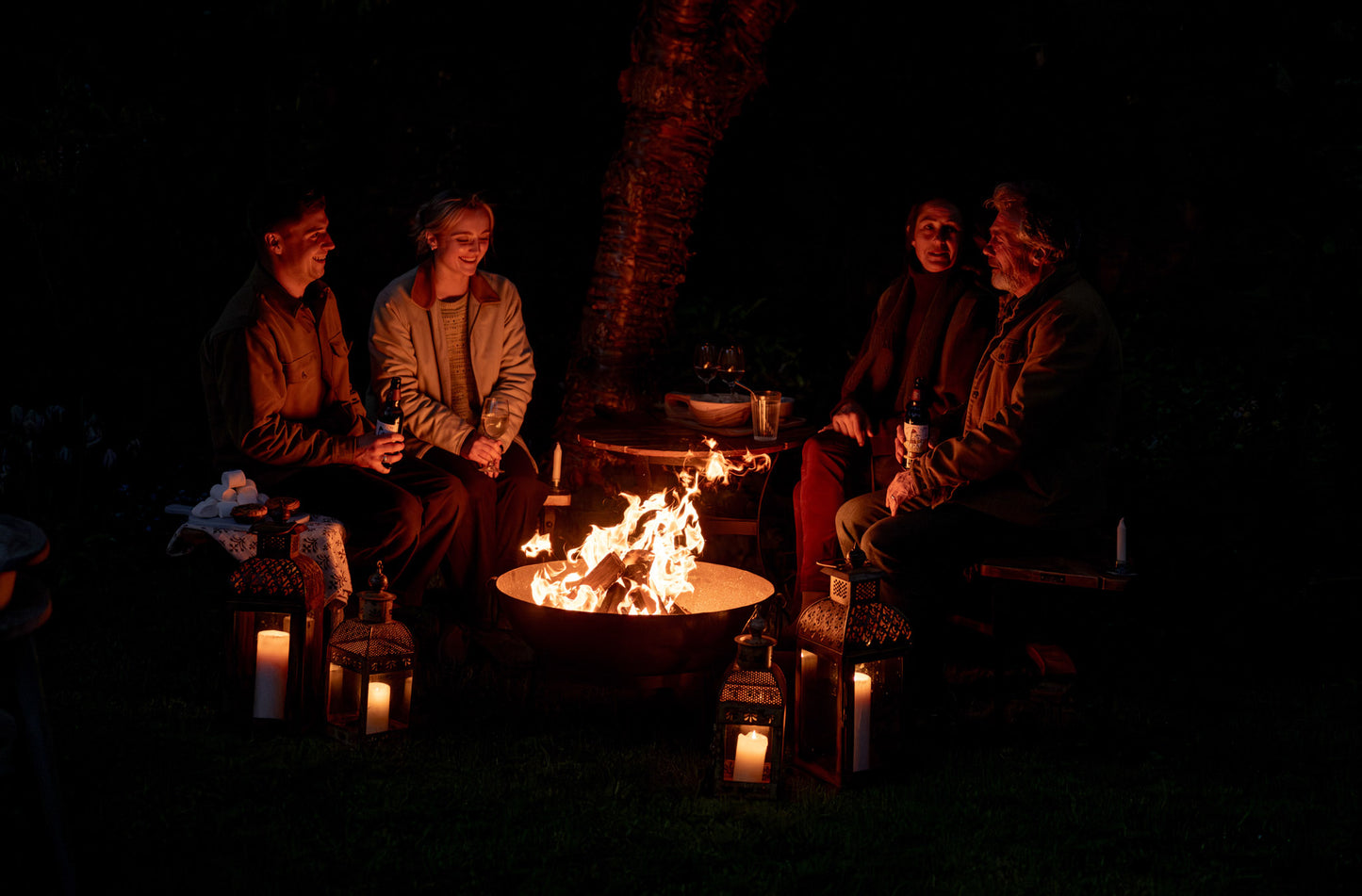Friends and family gathered around a glowing Kadai firebowl at night, enjoying drinks and conversation beside open flames and lantern light outdoors