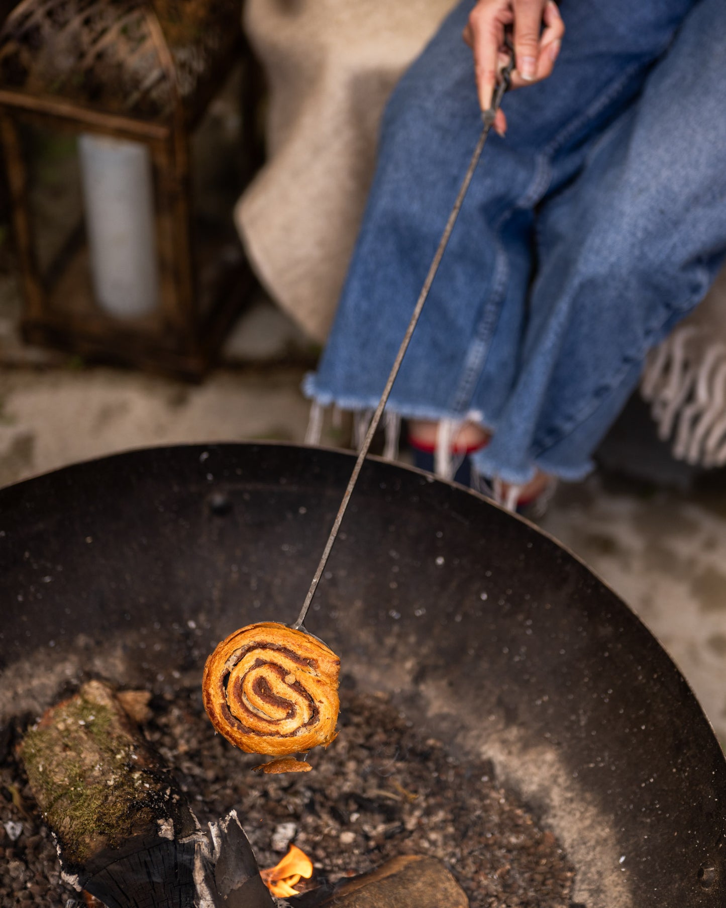 Person roasting a spiral pastry over an open fire pit using a long skewer and large kadai cooking utensil.