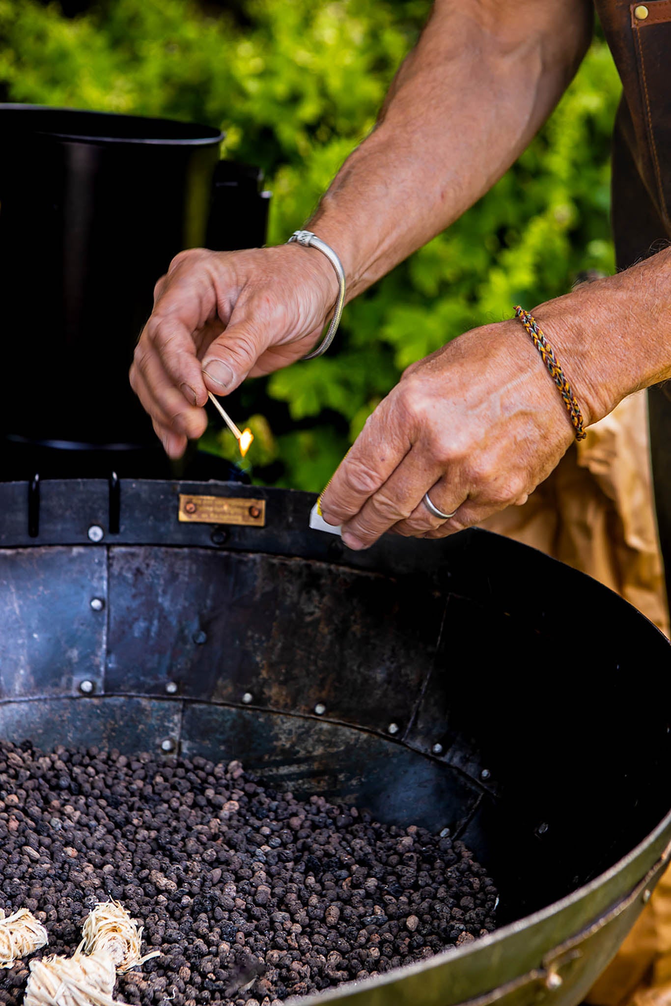 Person lighting charcoal in a barbecue grill with greenery in the background
