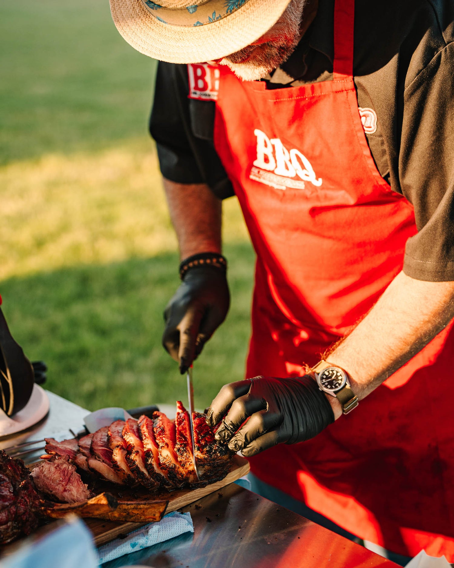 Person in a red apron with 'BBQ' on it cutting meat outdoors.