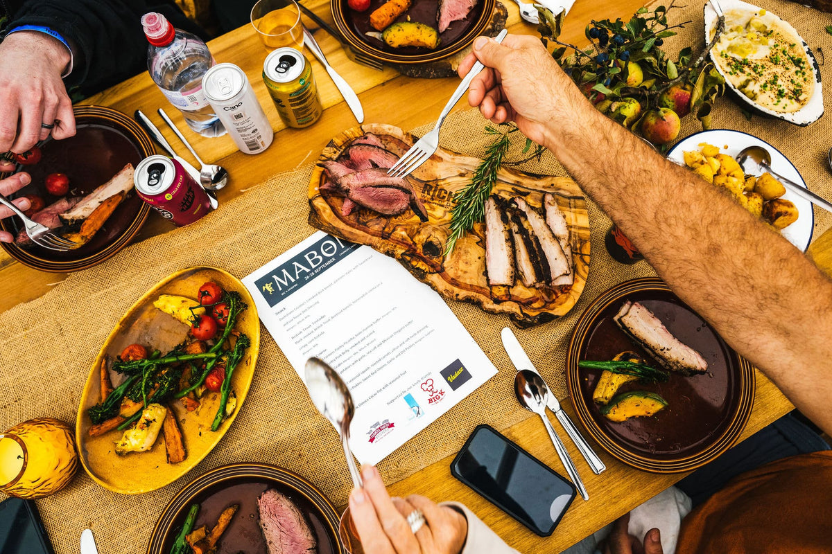 People eating on a table placing various meats and vegetables onto plates. a White menu named 'Mabon' sits on the table.