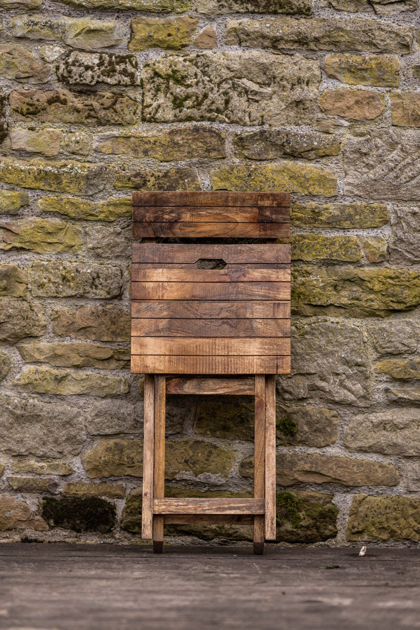 Folded rustic wooden café chair standing against a textured stone wall, showcasing its compact space-saving design