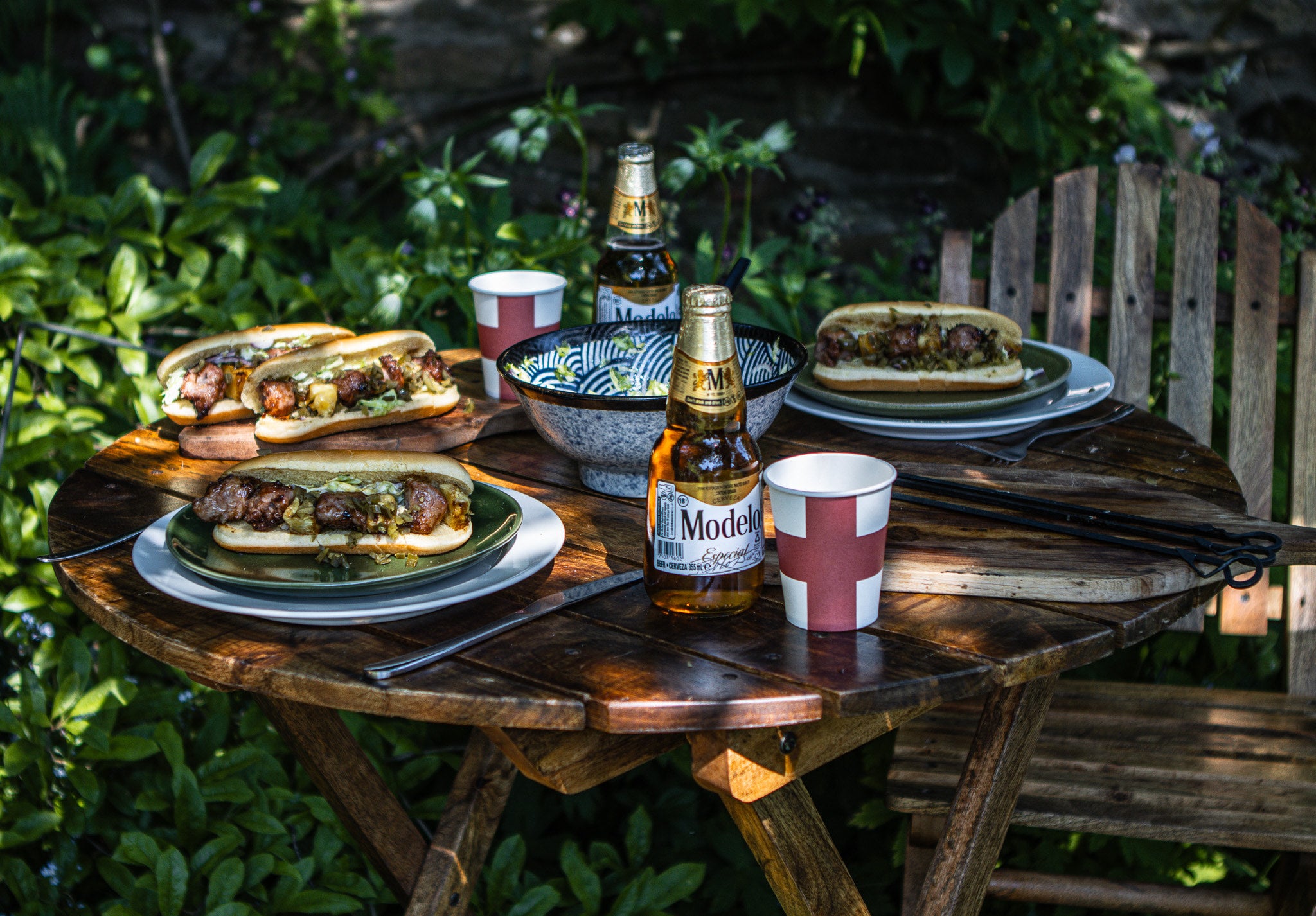 Outdoor setting with hot dogs on plates, beer bottles, and cups on a wooden table.
