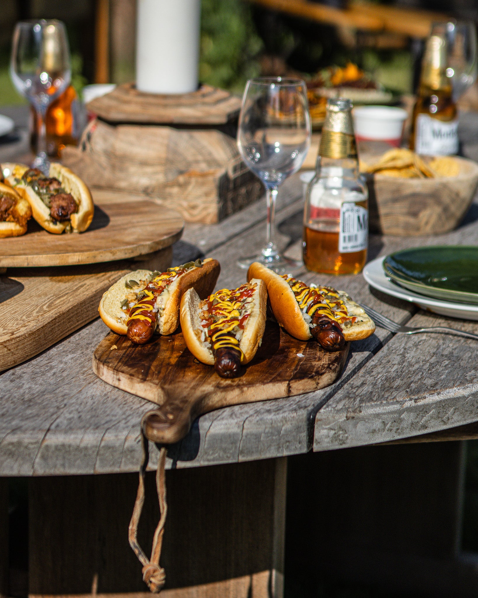Hot dogs with mustard on a wooden cutting board on a rustic outdoor table with beer and glasses.