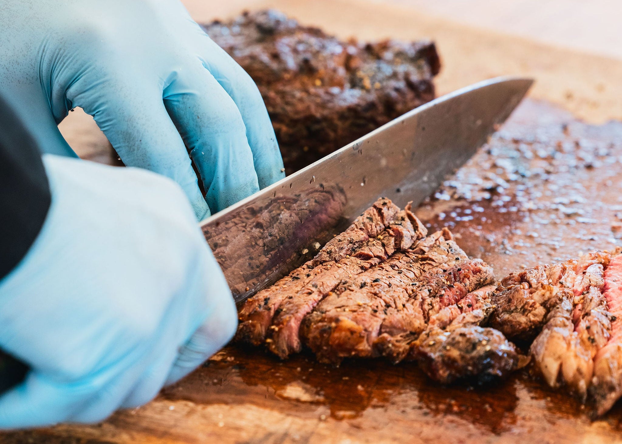 Person cutting meat on a wooden board with a knife, wearing blue gloves.