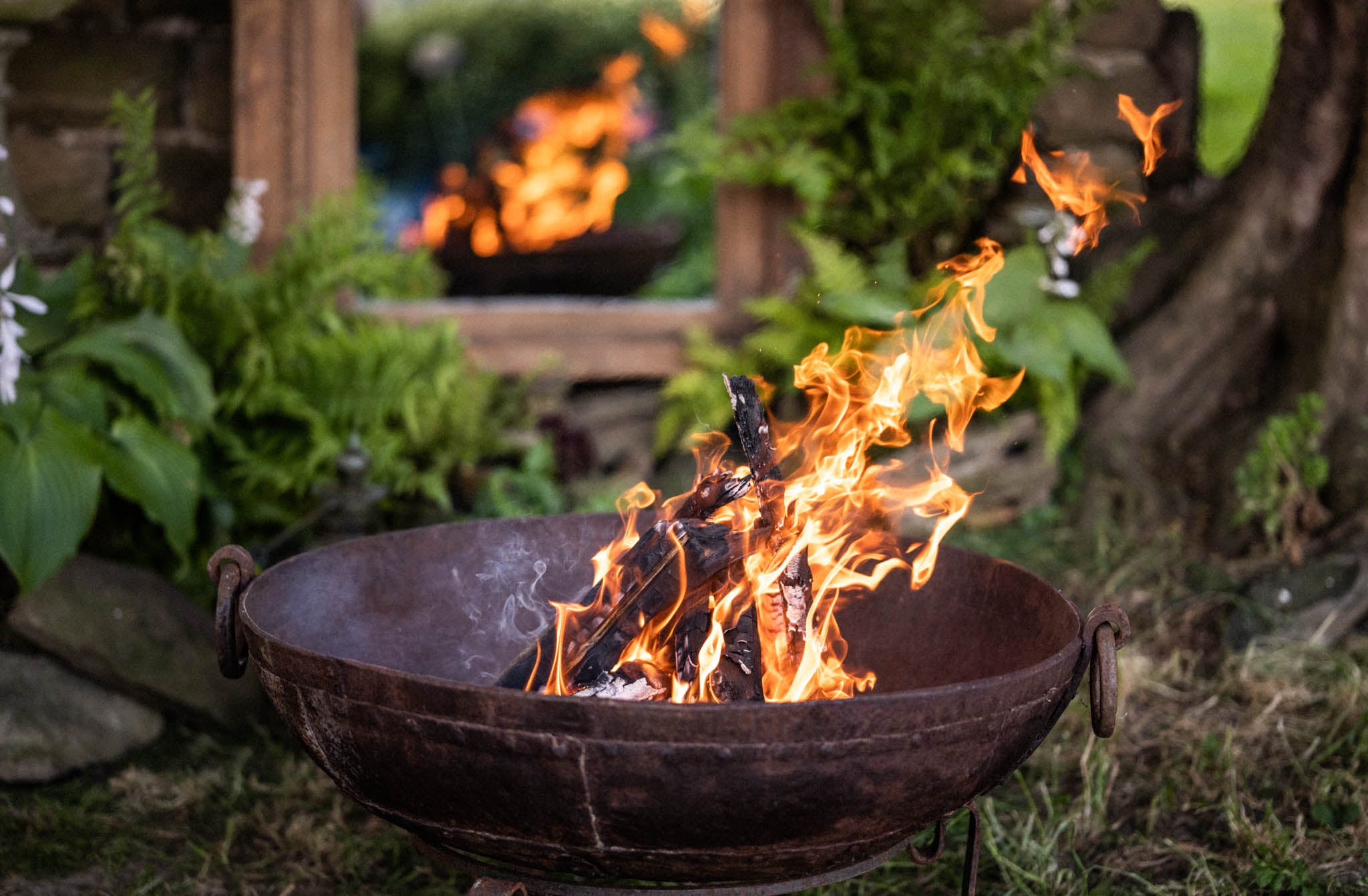 Fire burning in a metal Kadai bowl outdoors with greenery and a wooden bench in the background