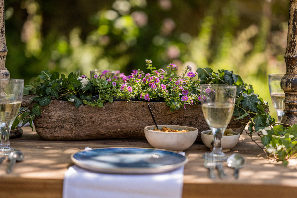 Outdoor setting with a table set for a meal, featuring a wooden planter with flowers and bowls.