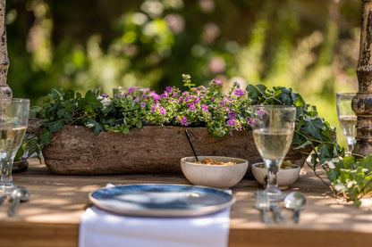 Outdoor setting with a table set for a meal, featuring a wooden planter with flowers and bowls.