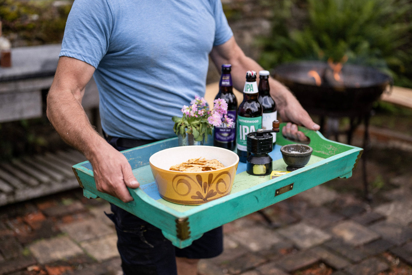 Person carrying a turquoise folding tray table with drinks, snacks and flowers beside an outdoor fire bowl on a rustic patio.