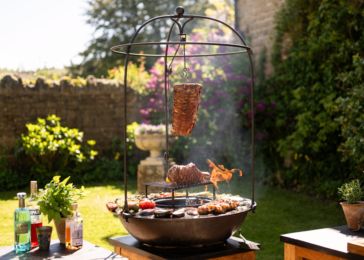 Kadai BBQ Plancha with suspended live-fire cooking, featuring open flame grilling, hanging meat, chargrilled vegetables and authentic outdoor barbecue entertaining in the garden