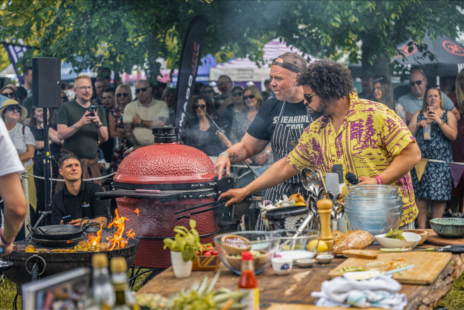 Two chefs cooking at a food festival with a large grill and ingredients on a table.