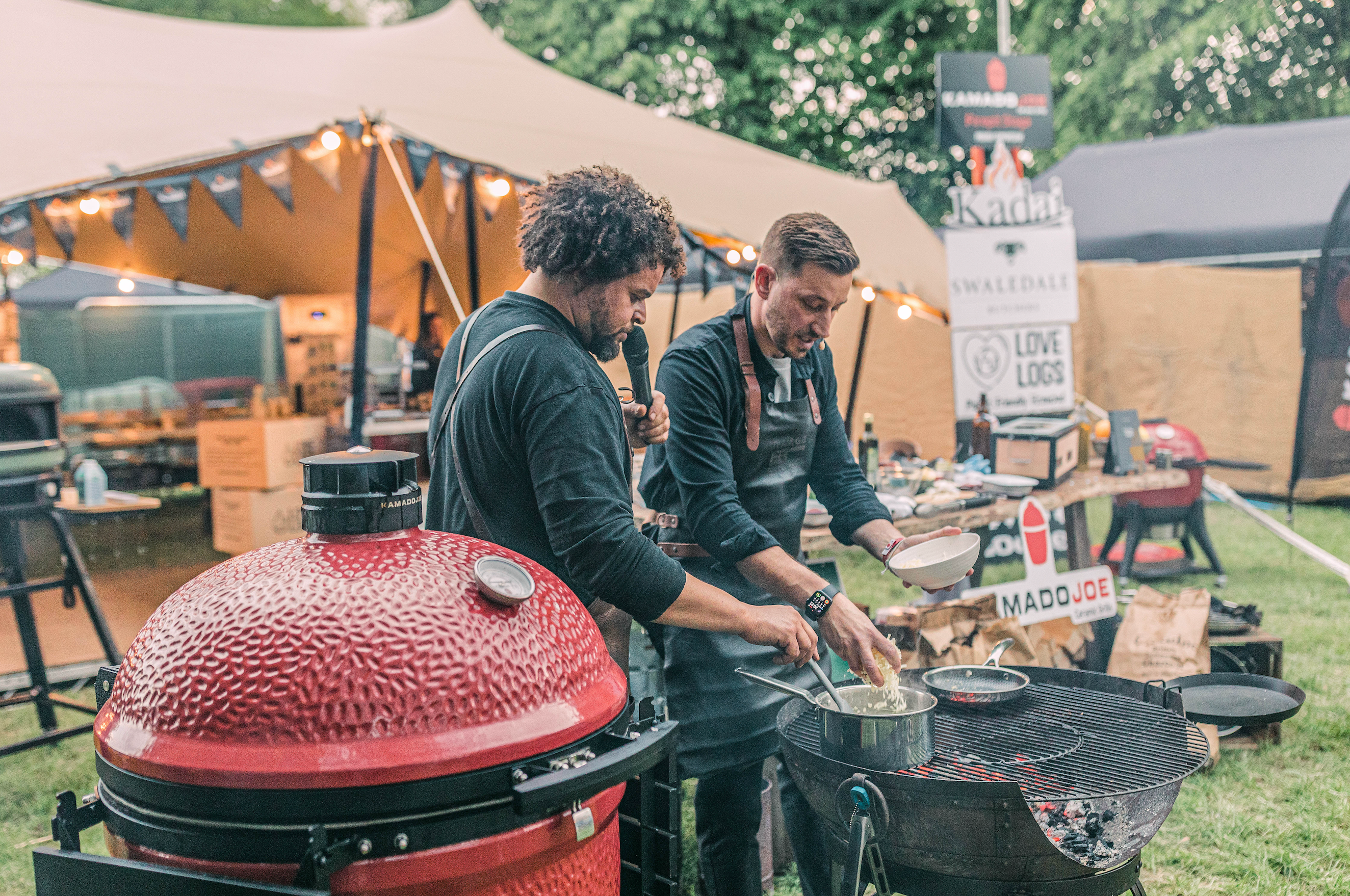 Two men cooking outdoors at a food festival with a large red barbecue and tents in the background.
