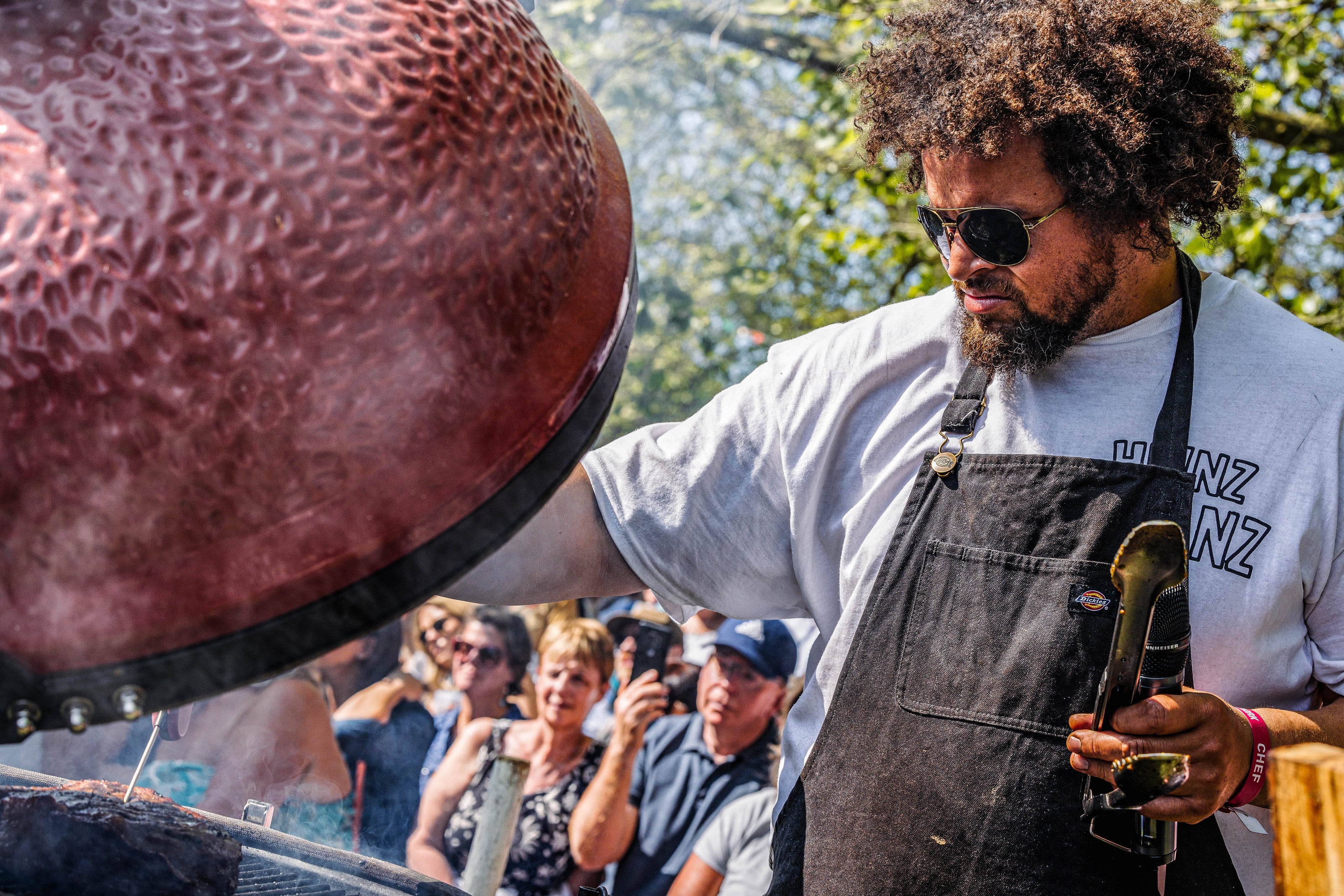 Person wearing an apron and sunglasses, standing in front of a large red object with people in the background.