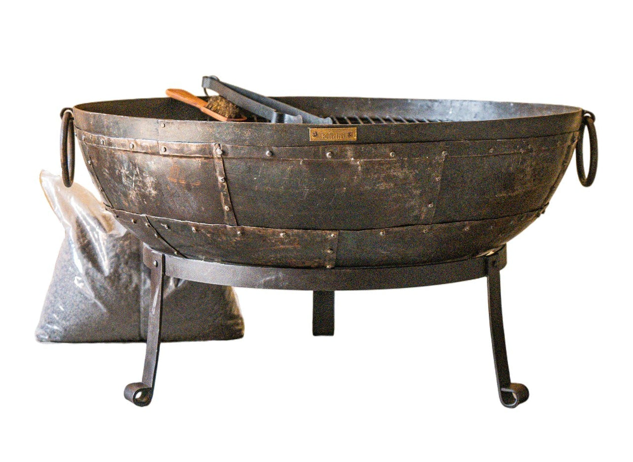 A round black metal fire pit with a stand and hanging side handles, shown on a plain white background. A a pair of tongs rest inside the pit.