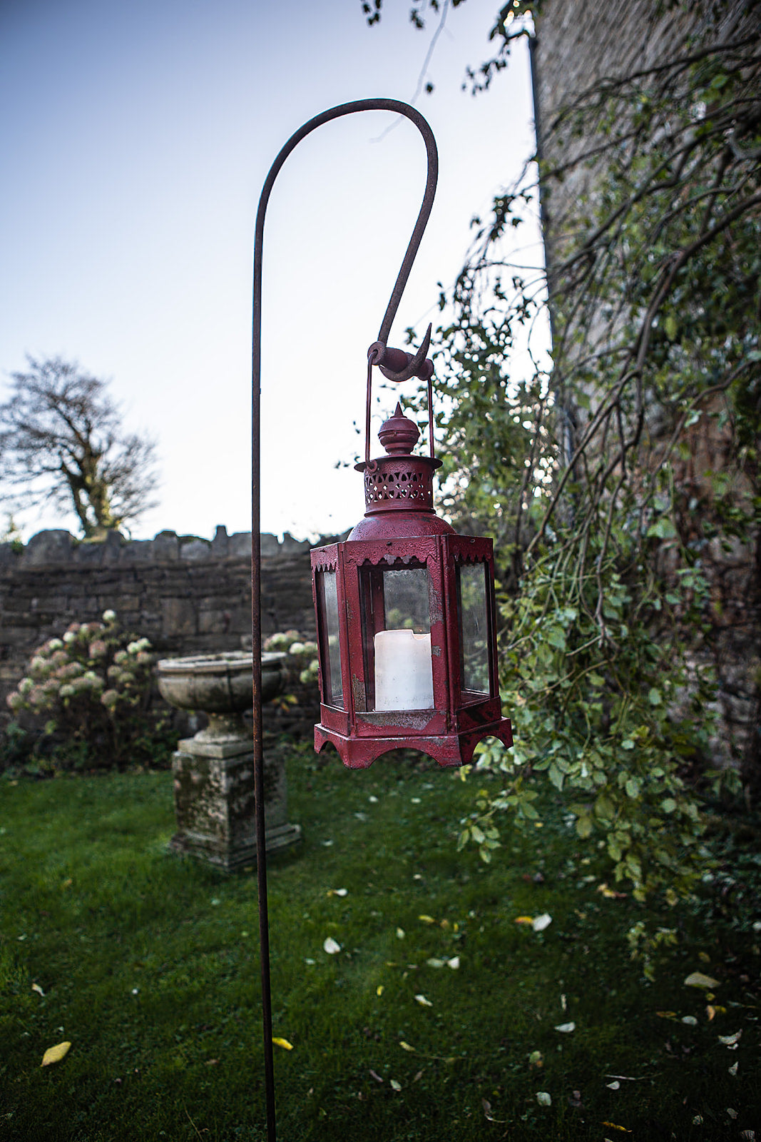 Red lantern hanging outdoors on a garden path with a stone wall and trees in the background.