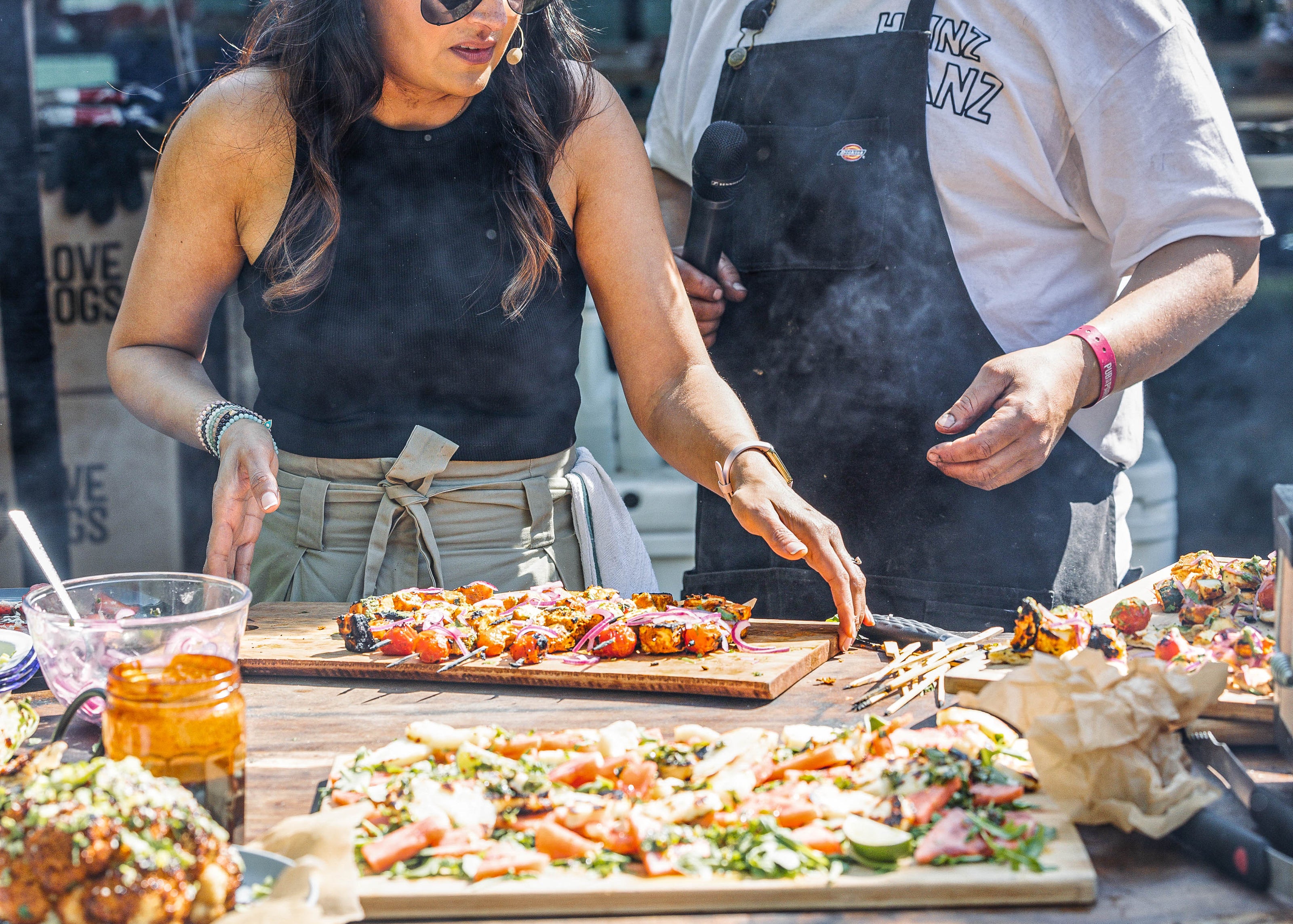 Two people preparing skewers at an outdoor event with a food truck in the background.