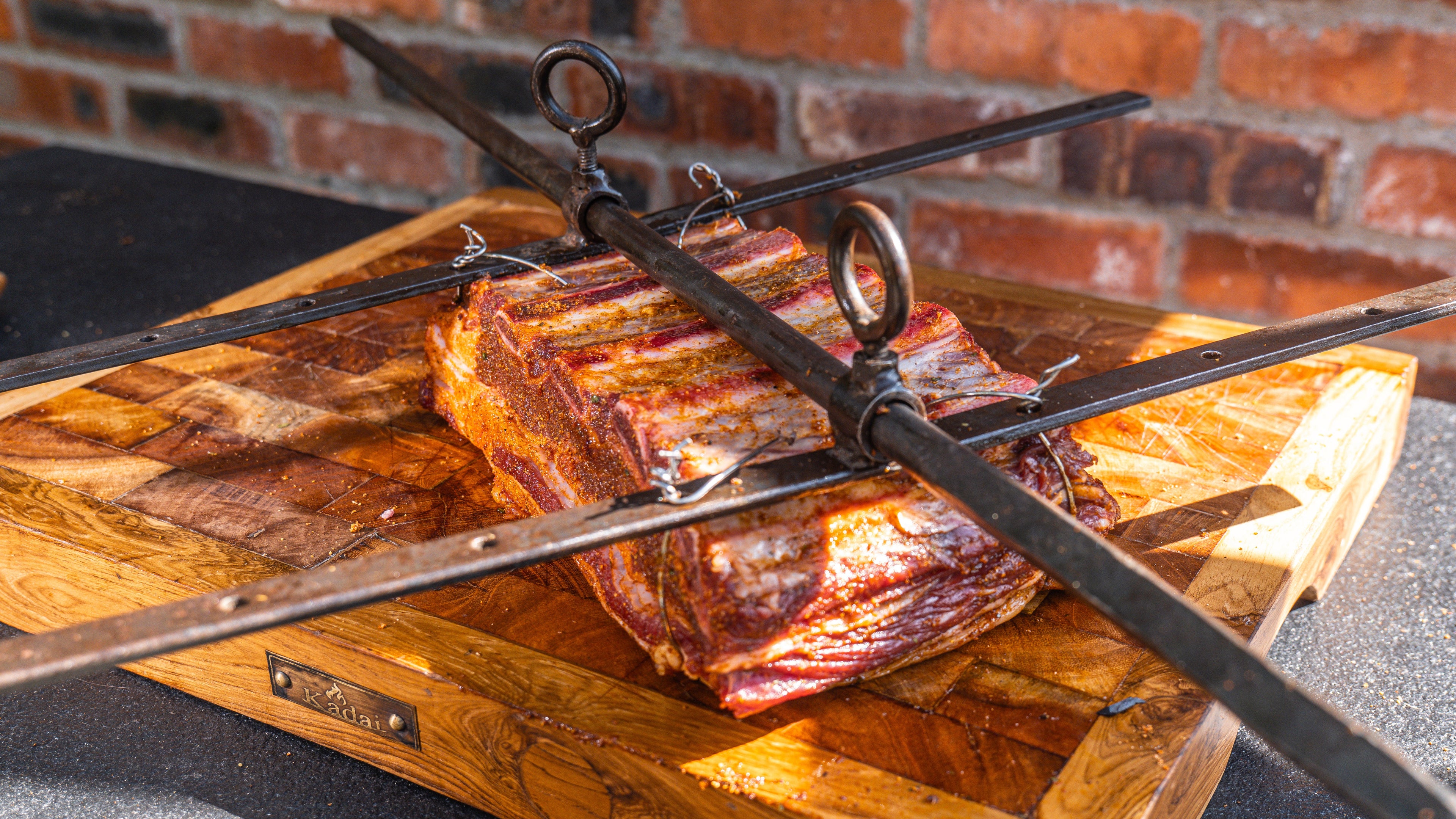 Wooden cutting board with ribs and a metal meat hook against a brick wall.