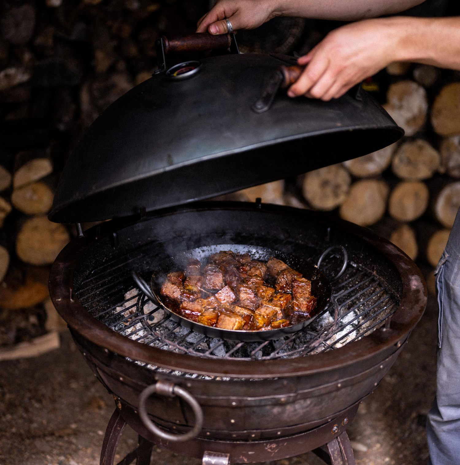 Person opening the smoker lid of a Kadai charcoal grill with food cooking inside, surrounded by stacked logs.