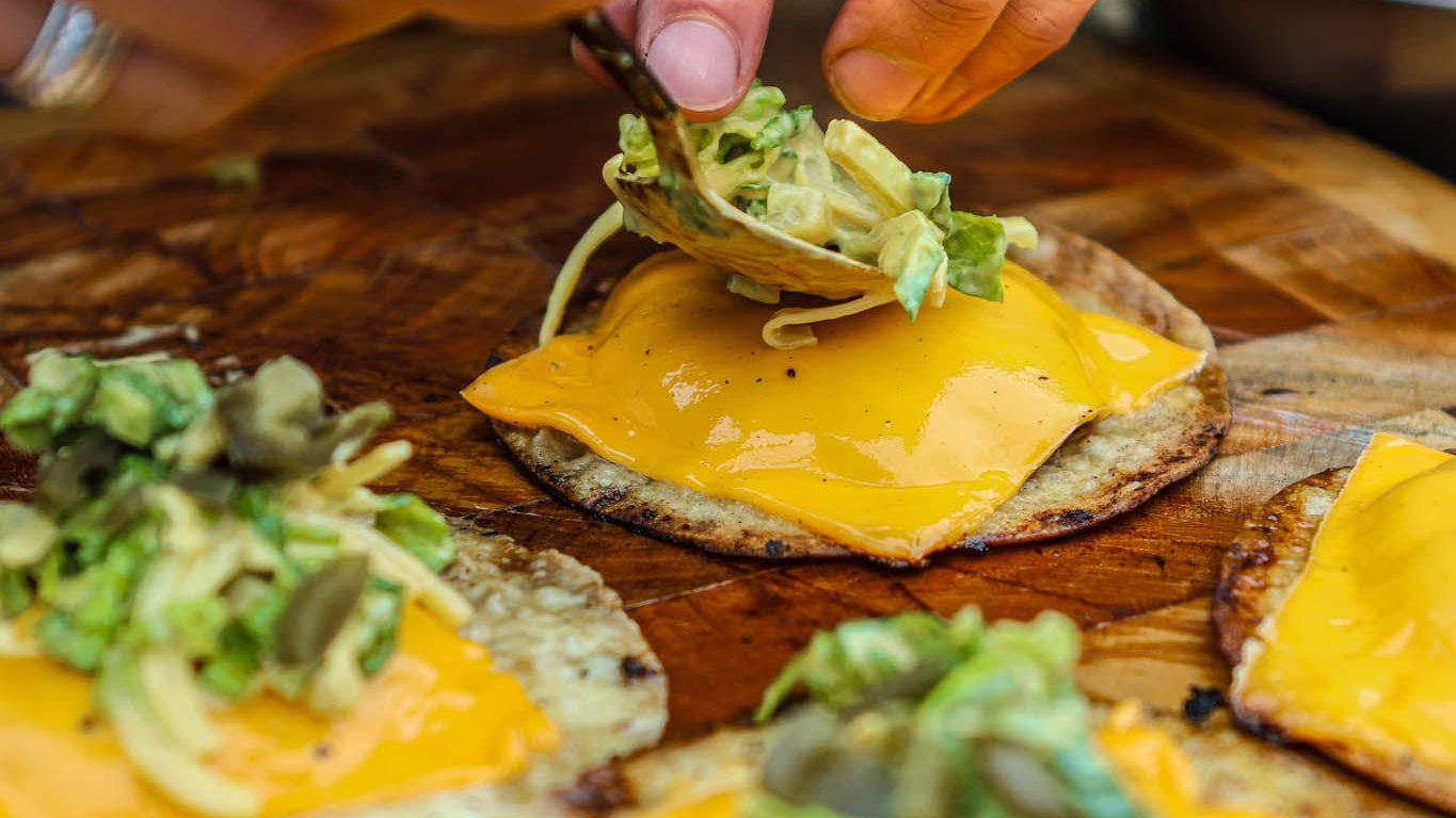 Person preparing food on a wooden cutting board with cheese and green toppings.