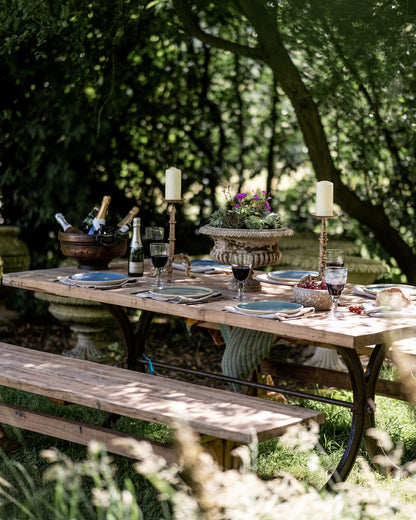 Rustic teak garden bench paired with a long outdoor dining table set with wine, candles and tableware beneath shaded trees in a garden setting.
