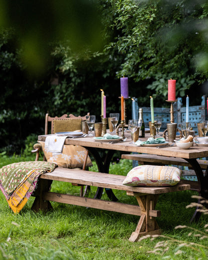 Rustic teak garden bench beside a long outdoor dining table set with colourful candles, cushions and tableware in a lush garden setting.