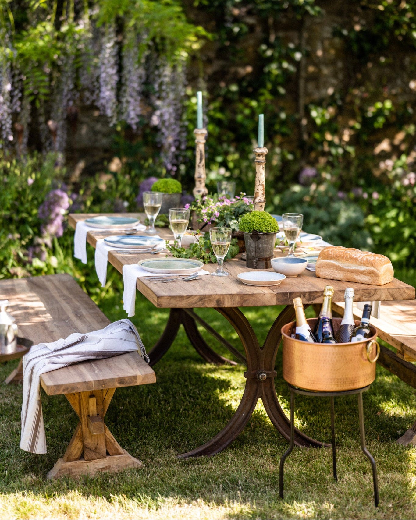 Outdoor dining setup with a wooden teak garden table, benches, and a bottle of wine in a garden.
