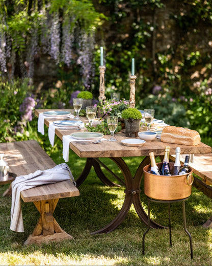 Outdoor dining setup with a wooden teak garden table, benches, and a bottle of wine in a garden.