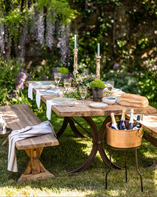 Outdoor dining setup with a wooden teak garden table, benches, and a bottle of wine in a garden.