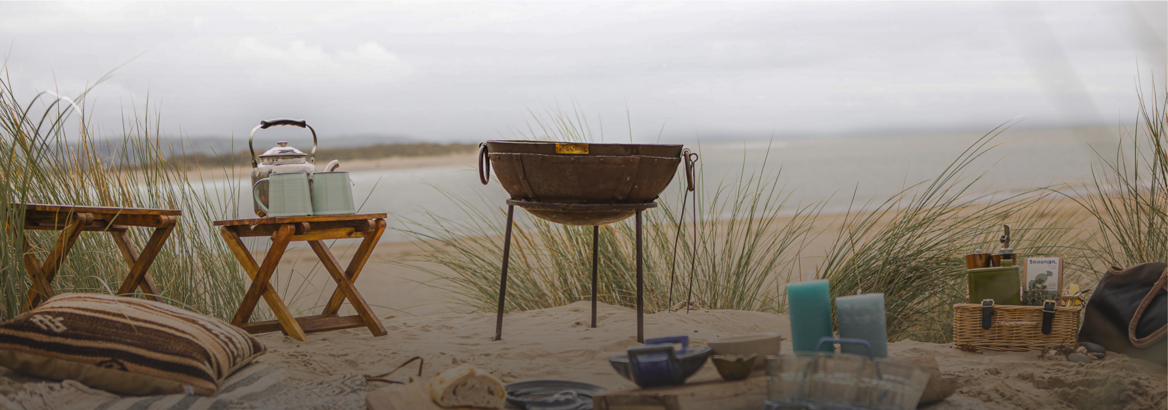 Outdoor setting with a camping grill, cooking accessories, tables, and chairs on a sandy beach.
