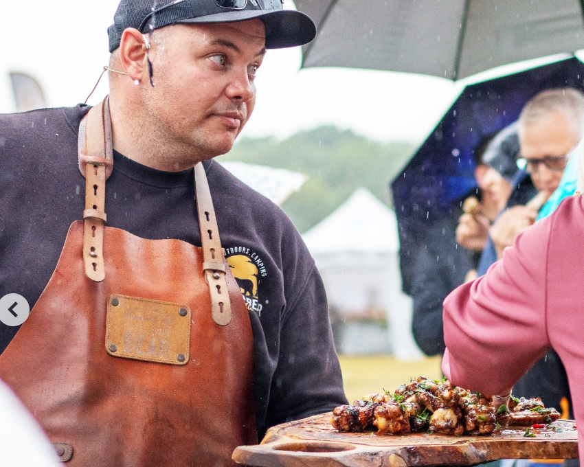 Man in a leather apron holding a tray of food outdoors with people and umbrellas in the background