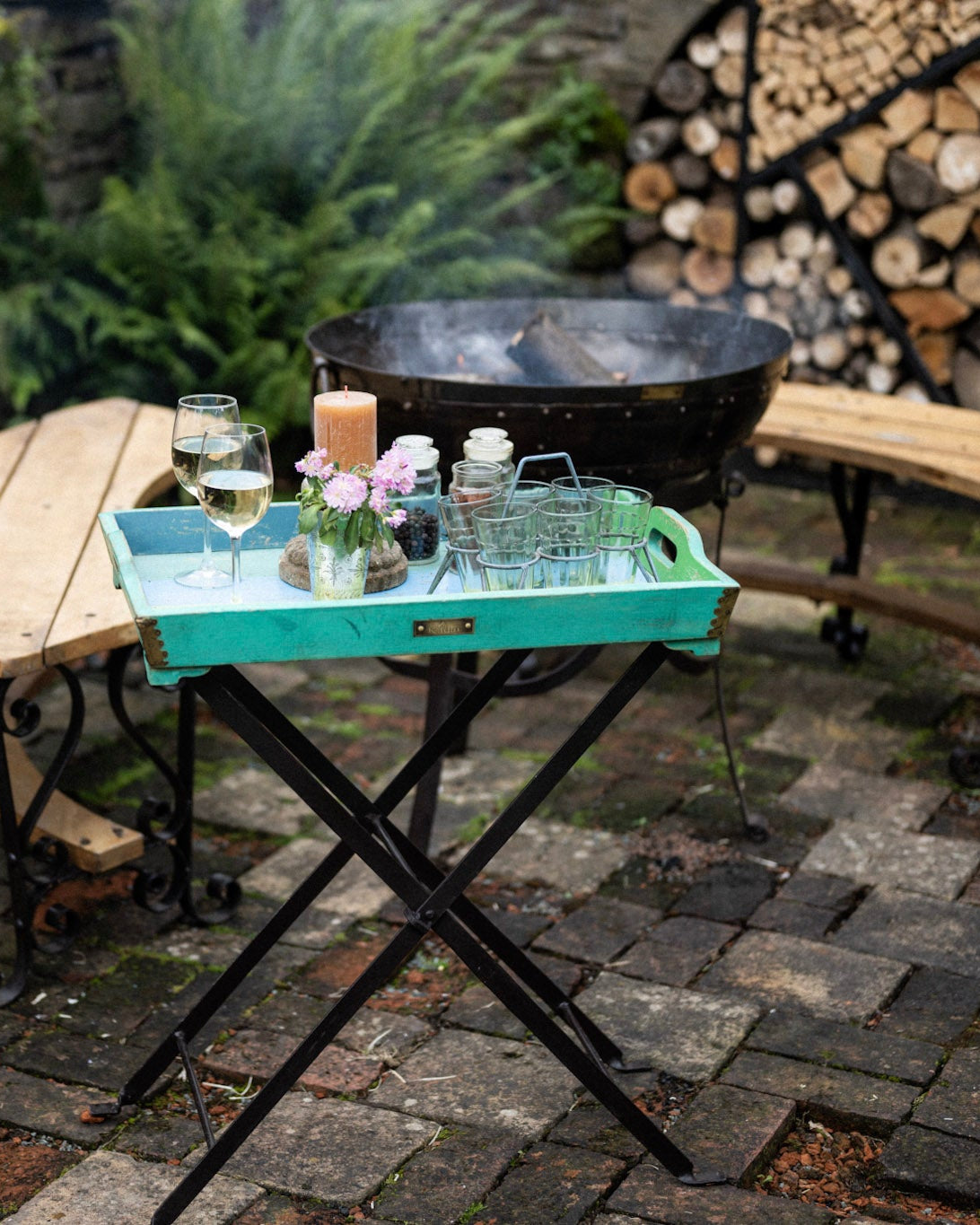 Turquoise serving tray on a folding stand with glasses, flowers and candles beside a rustic outdoor fire bowl and wooden seating area.