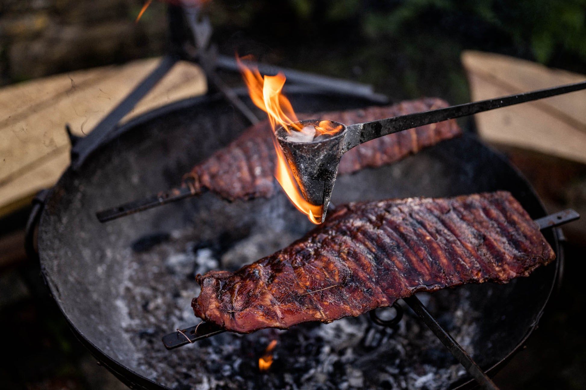 Racks of ribs cooking on an asado cross while a flaming flambadou seasons the meat over open coals